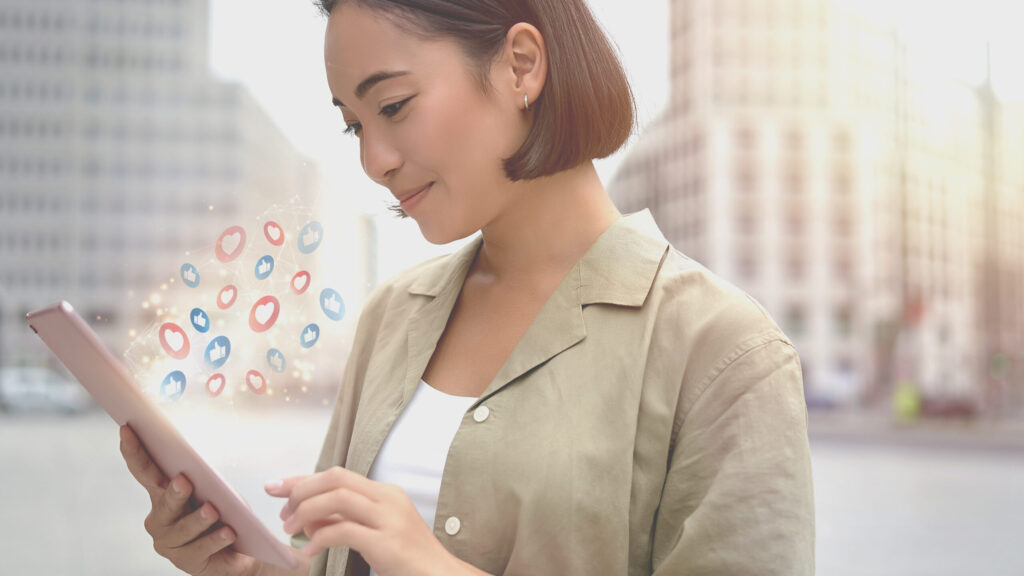 Oriental woman looking at a tablet while smiling. Social media icons appear on the screen. In the background, a city.