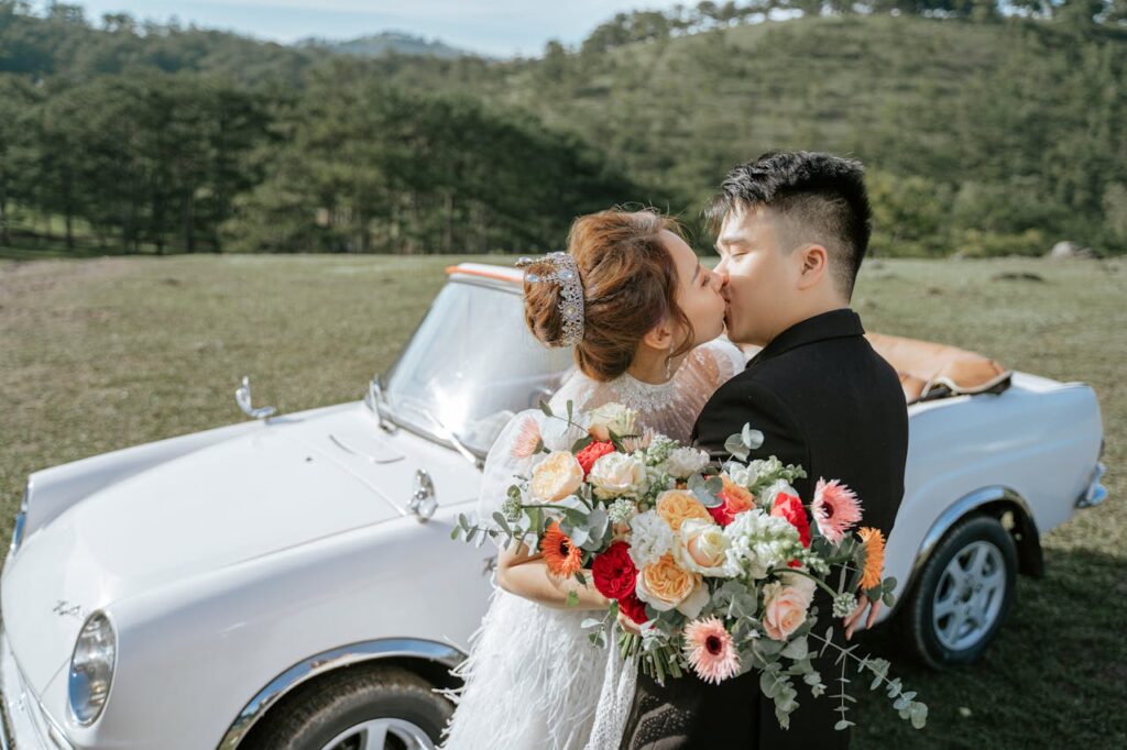 Oriental couple dressed as bride and groom kissing. They are in front of a convertible car. In the background, mountains.